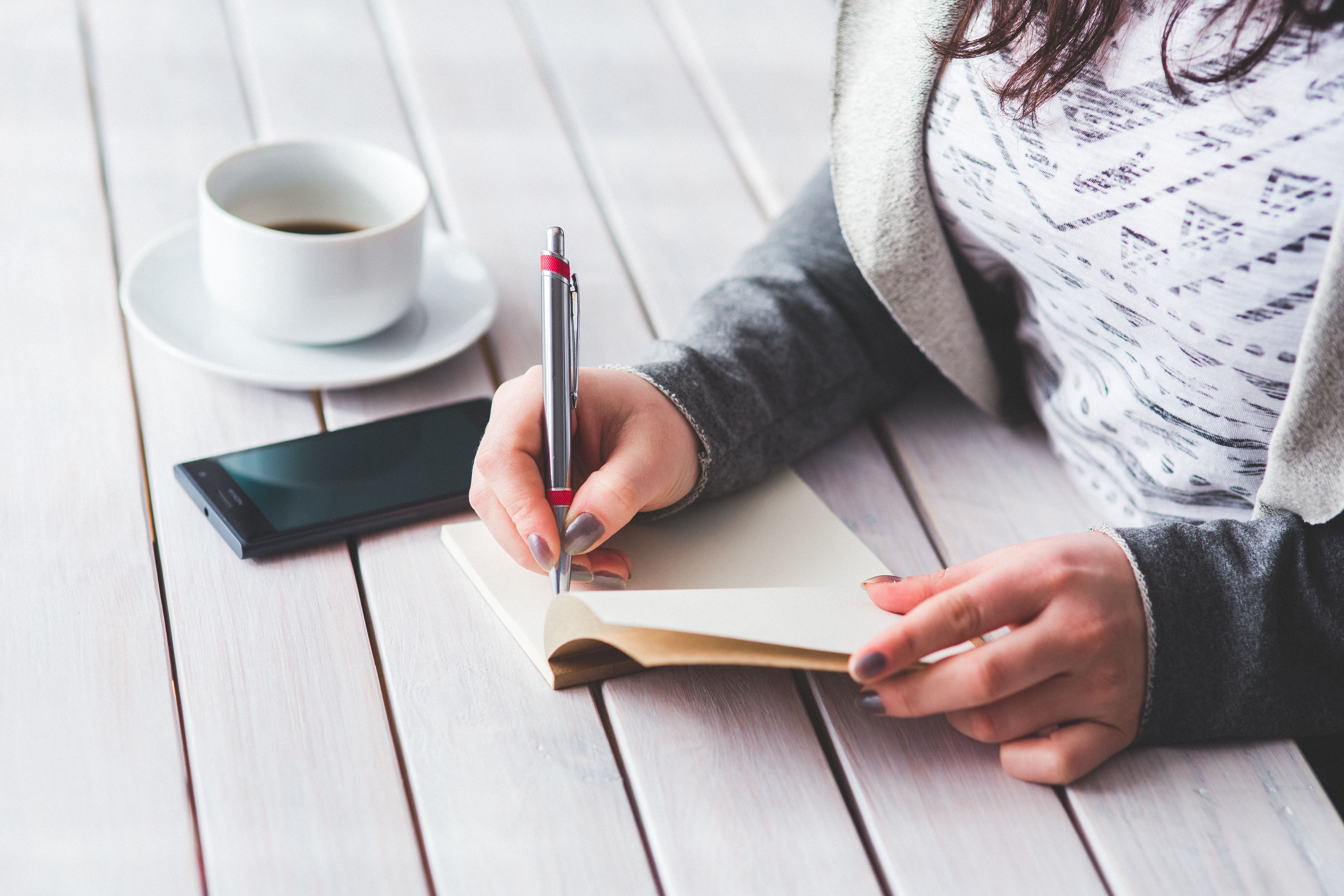 Person writing in a notebook beside a tablet and cup of coffee.
