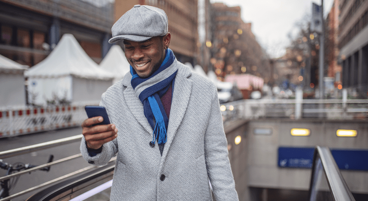 A photo of a man outdoors, walking in an urban setting. He is dressed in a coat and scarf, holding a smartphone and smiling.