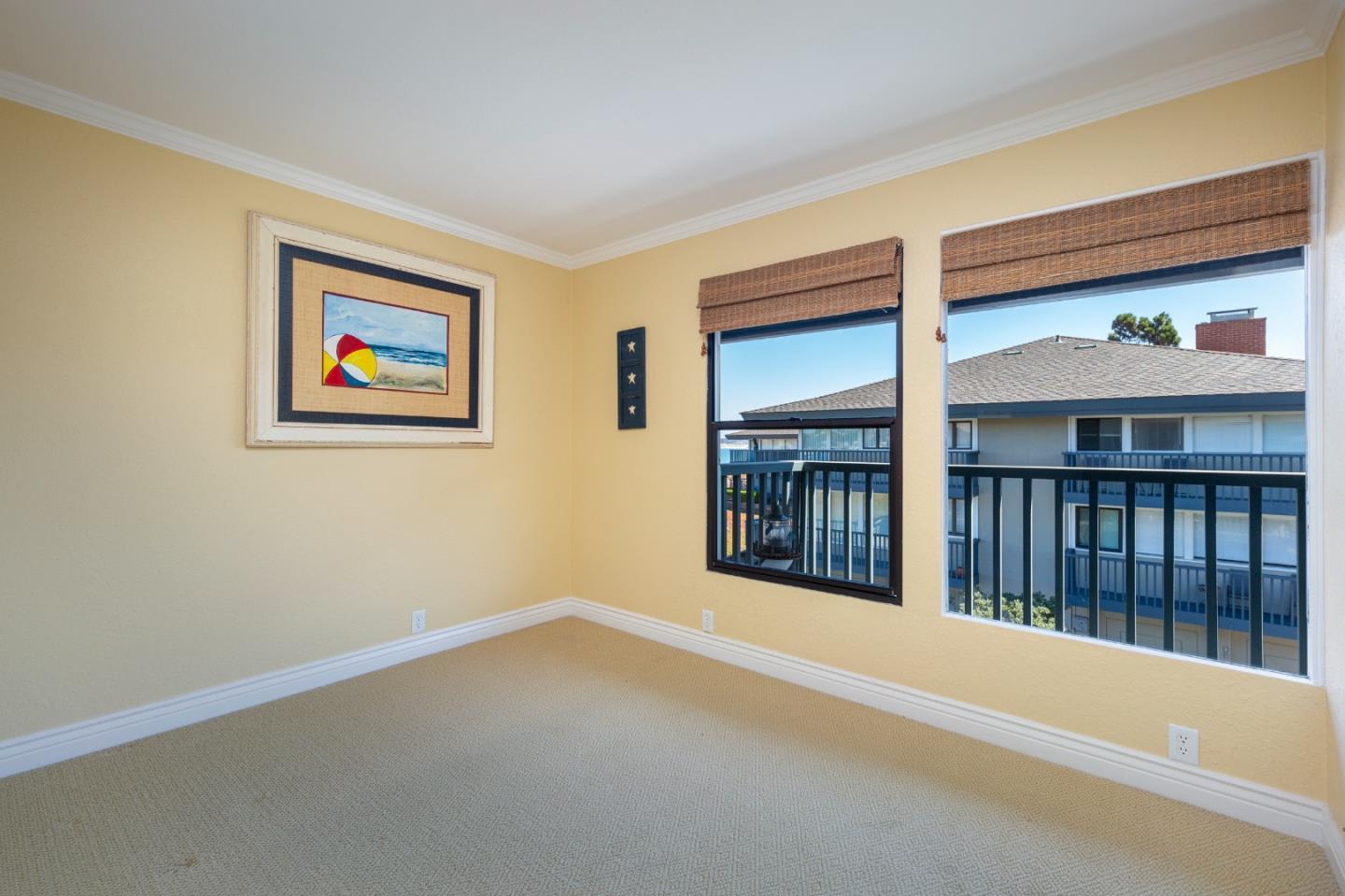 Unfurnished room with beige carpet, woven window shades, and framed beach artwork.