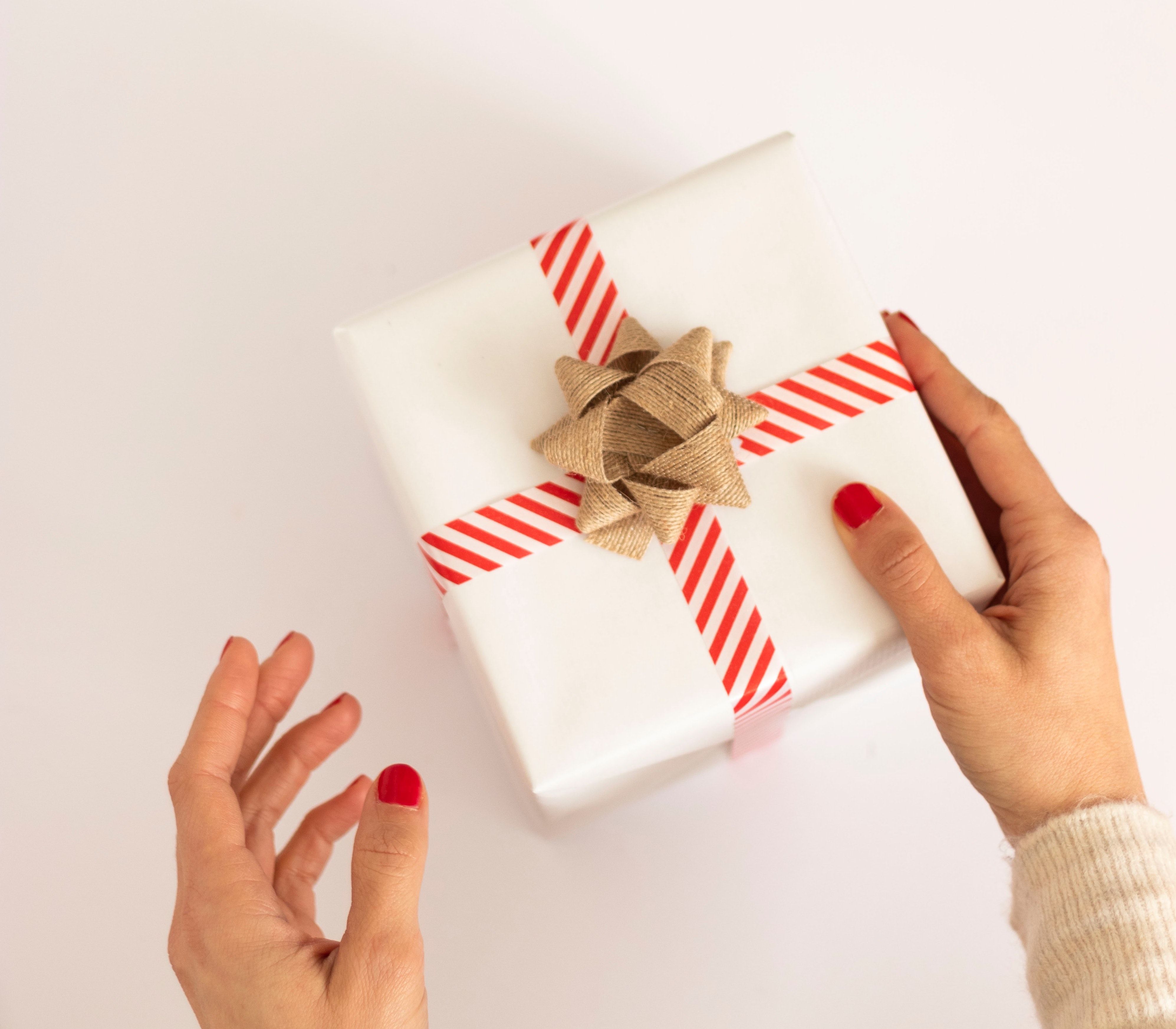 Hands pass a white gift box with red striped ribbon and a burlap bow.
