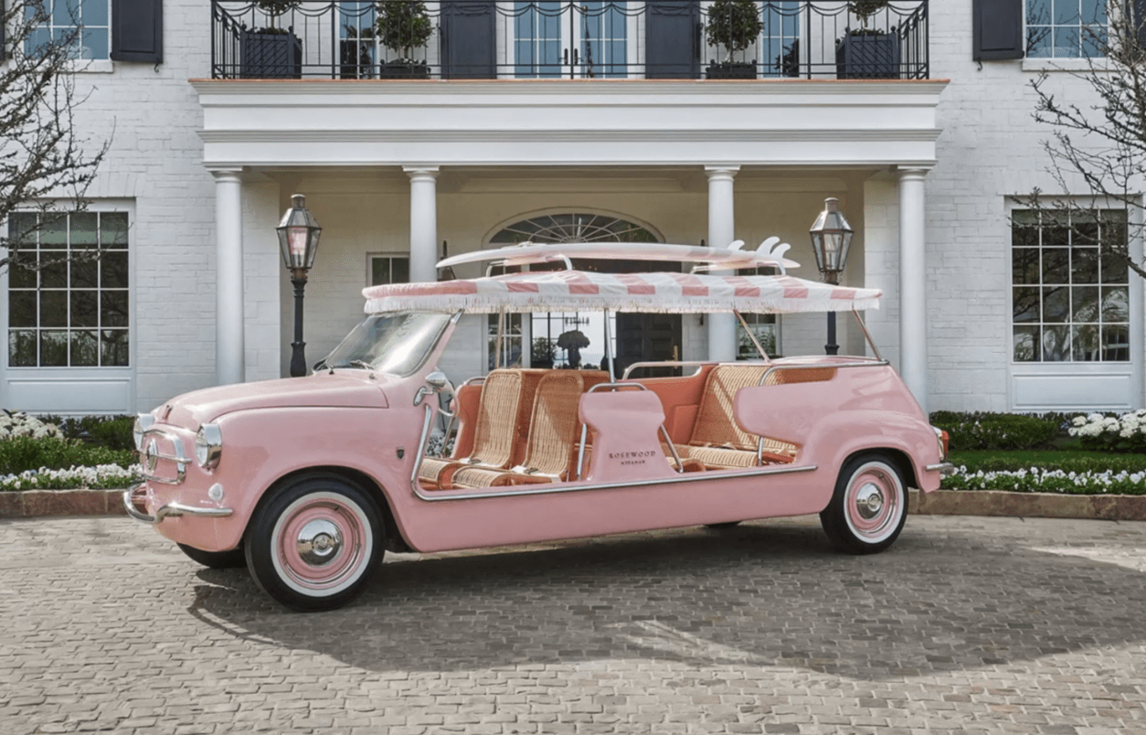 A pink car with a surfboard rack sits in front of a white mansion.