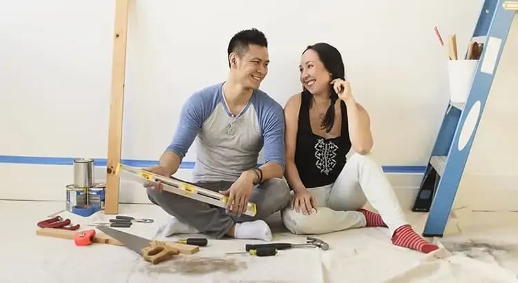 A couple sitting on the floor, surrounded by painting supplies and decor items. They are smiling and appear to be in the process of renovating or decorating their home.