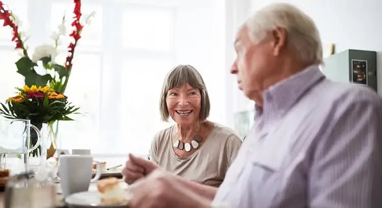 An elderly couple enjoying a meal together at a dining table. They are smiling and seem to be having a pleasant conversation, suggesting themes of companionship and happiness in later life.