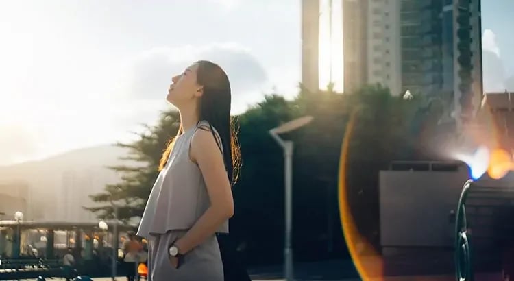 A woman standing outside in a city setting, basking in the sunlight. She appears to be enjoying the moment, possibly indicating freedom, joy, or taking a break.
