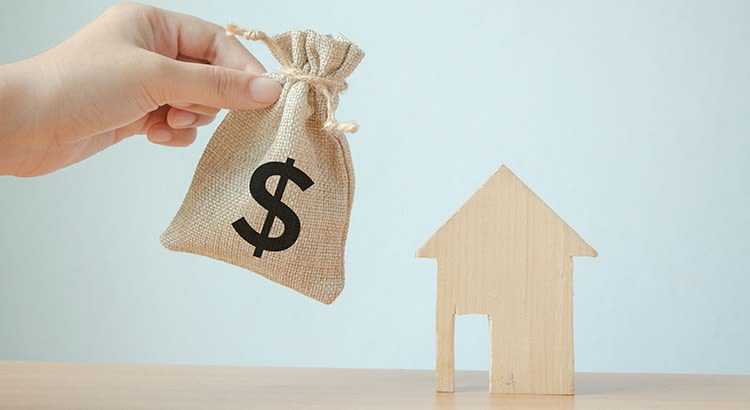  A small bag with a dollar sign being placed next to a wooden house model.