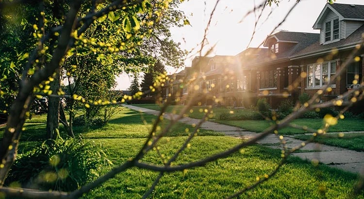 A photograph of a suburban neighborhood during sunset, with houses, trees, and a well-maintained lawn visible.