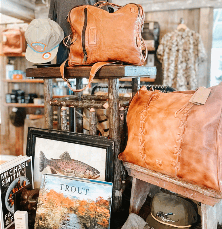 Rustic shop display of tan leather bags, a cap, and framed fishing artwork.