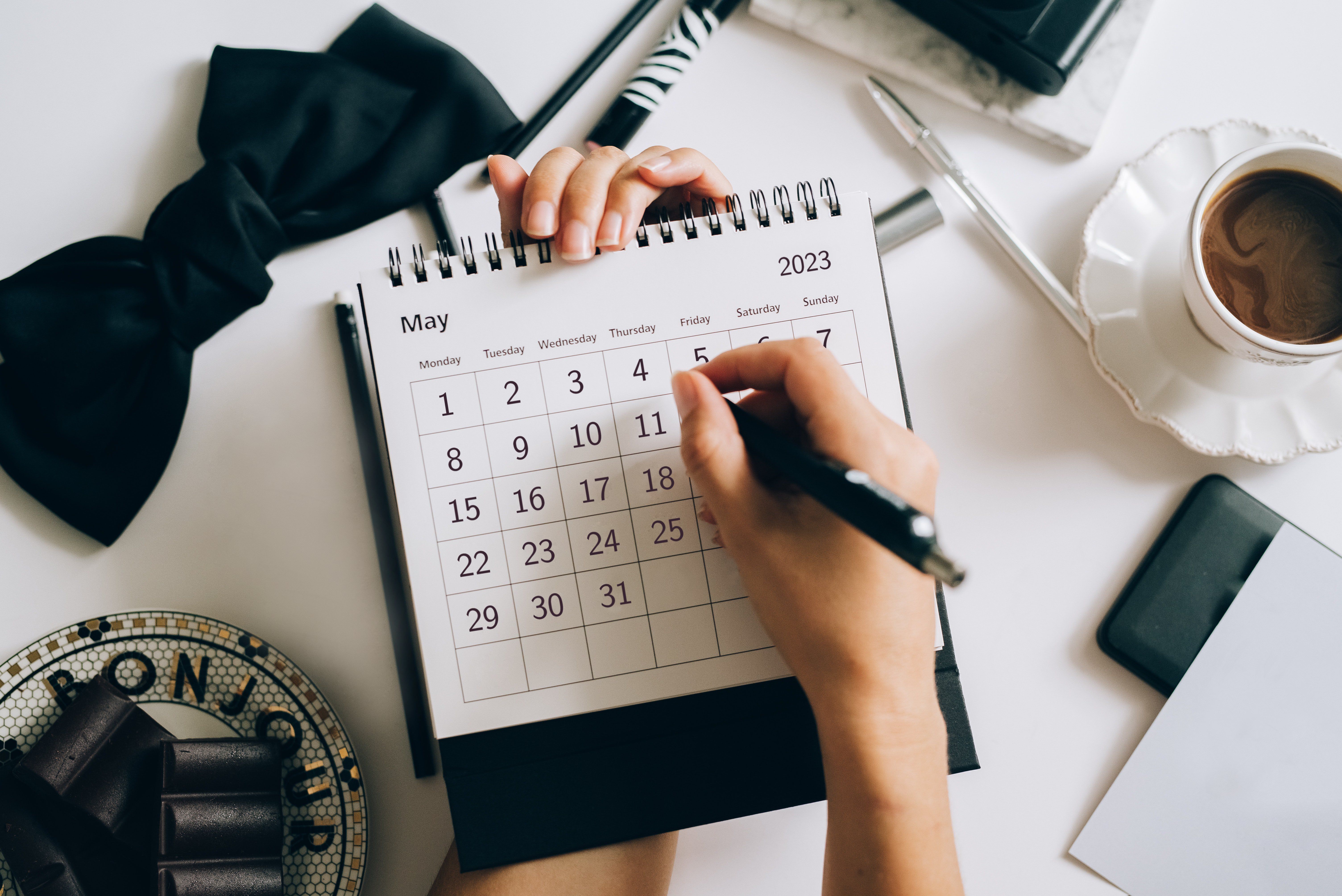 A hand marks a May 2023 calendar with a pen beside a cup of coffee.