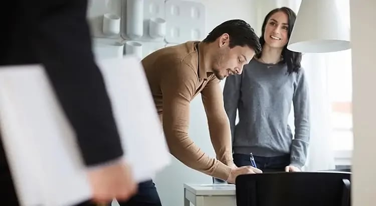 A man and a woman in an office setting, reviewing documents. The man is bending over a table, possibly signing papers, while the woman stands beside him, observing. The image suggests a professional or real estate transaction.