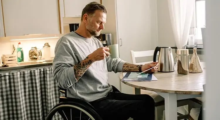 A man in a wheelchair is sitting at a kitchen table, holding a cup and looking at a tablet. The kitchen has a modern design with various kitchen appliances and utensils visible.