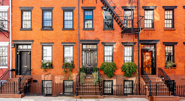 A row of brownstone buildings with a vibrant, urban aesthetic featuring a classic brownstone facades with red brick exteriors. Each building features several windows with black shutters, adding a contrasting color to the red bricks. The front doors are painted black and are framed by stone or brick.