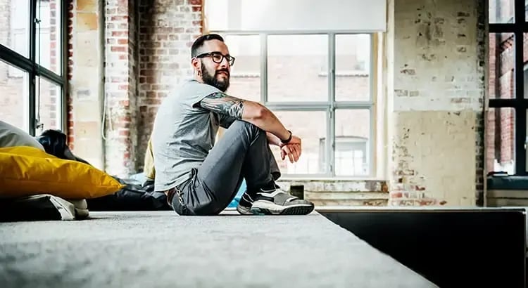  A man sitting on the floor next to a large window, looking relaxed and thoughtful. The setting is a modern, loft-style apartment with exposed brick walls.