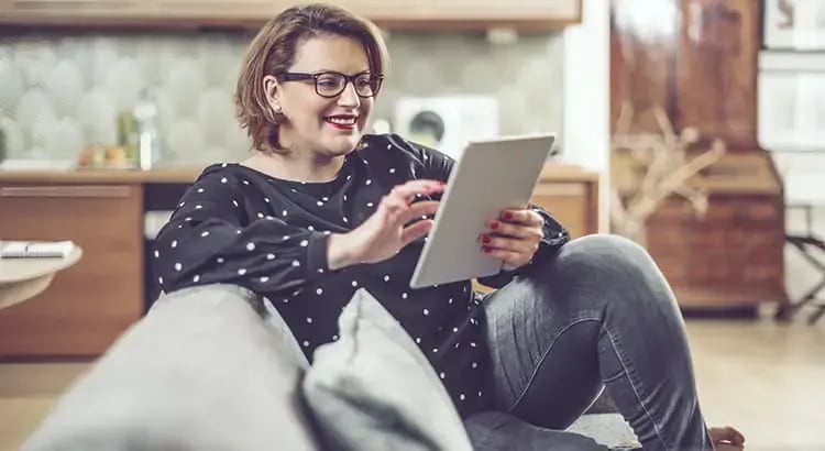 A woman sitting comfortably on a couch, holding and reading from a tablet. She is smiling, suggesting she is enjoying the content on the device.