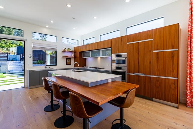Modern kitchen with warm wood cabinetry, a white island, and a wooden dining table beside large windows.