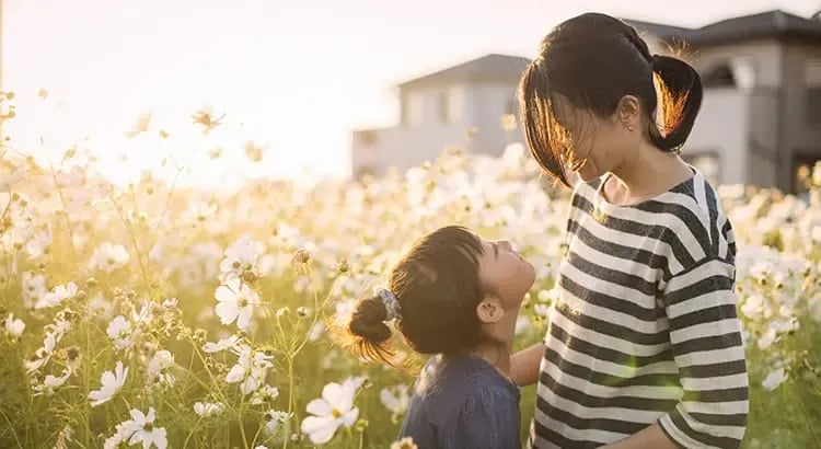 A mother and her young child, in a field of flowers, both smiling and enjoying the outdoor environment. The scene is bathed in warm, golden light.