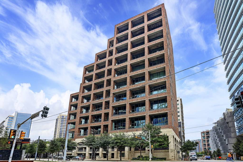 Brick residential tower with recessed balconies on a downtown street corner beneath a blue, cloud-streaked sky.