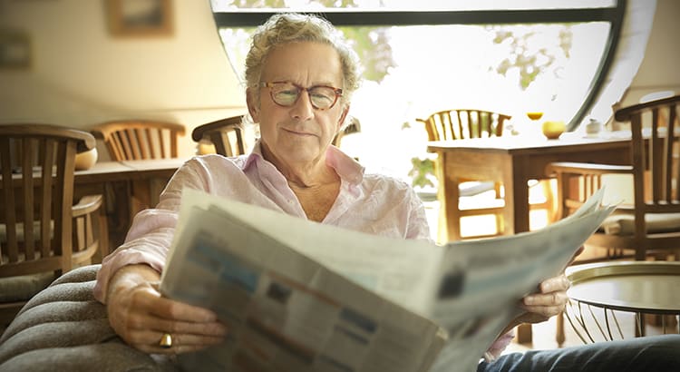 An older man sitting comfortably and reading a newspaper in a well-lit room. The setting suggests themes of staying informed, relaxation, and home comfort.