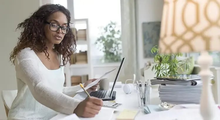 A woman with curly hair working at a table, using a tablet and surrounded by papers and plants.