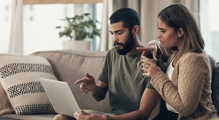 A couple sitting on a couch, looking at a laptop together. The man is typing on the laptop while the woman is holding a coffee cup and pointing at the screen.