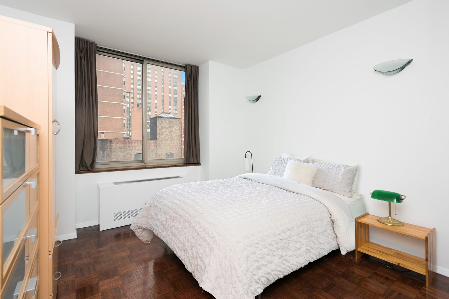 Bedroom with a white quilted bed, parquet floors, and a large window overlooking buildings.