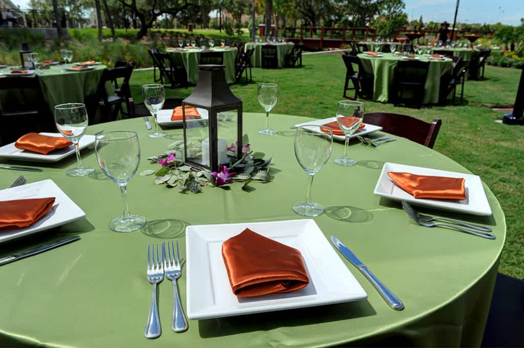 The image features an elegant outdoor event setup at Ulele, with beautifully arranged round tables adorned with green tablecloths and burnt orange napkins. Each table is accented with lantern centerpieces and pristine glassware, creating a charming and sophisticated ambiance for special occasions amidst a scenic backdrop.