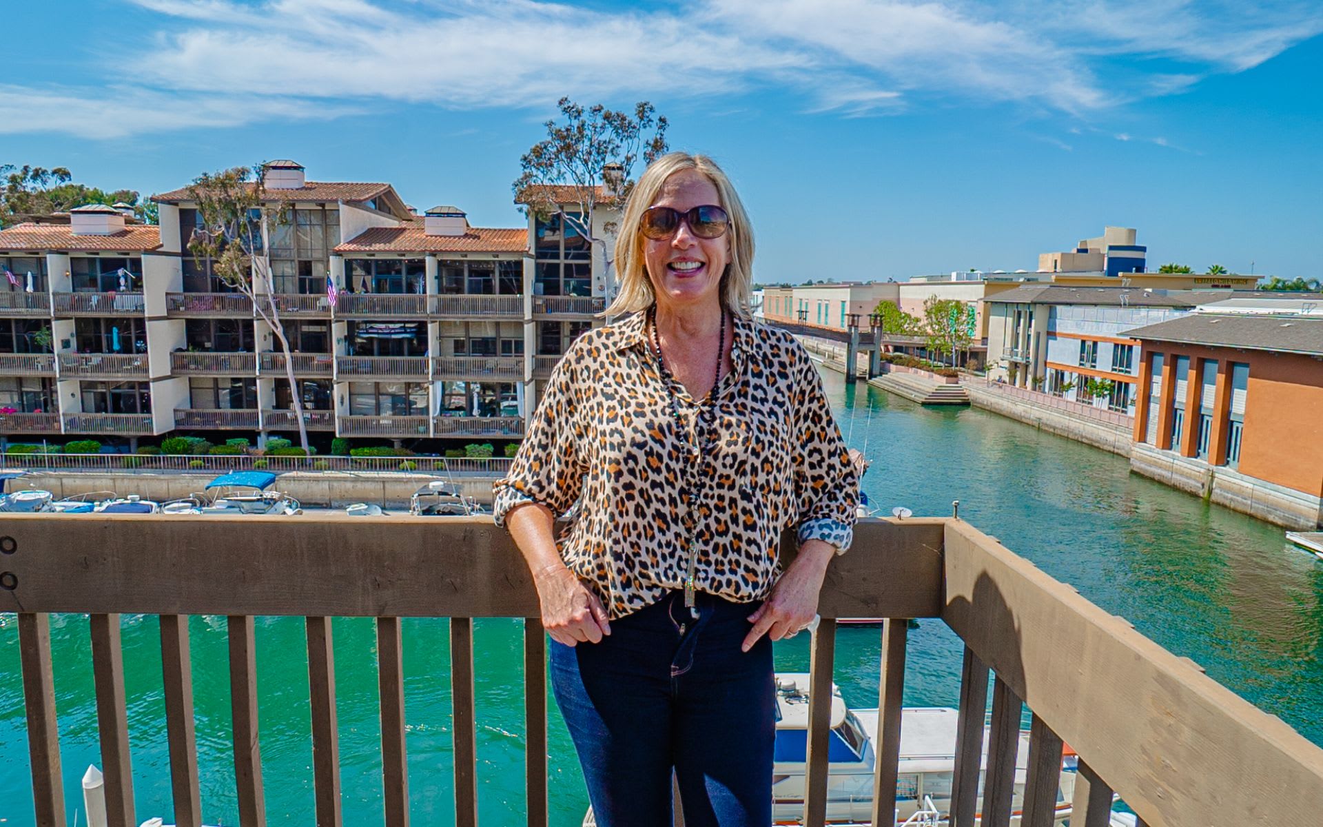 Woman in sunglasses and a leopard-print blouse on a balcony overlooking a marina canal.