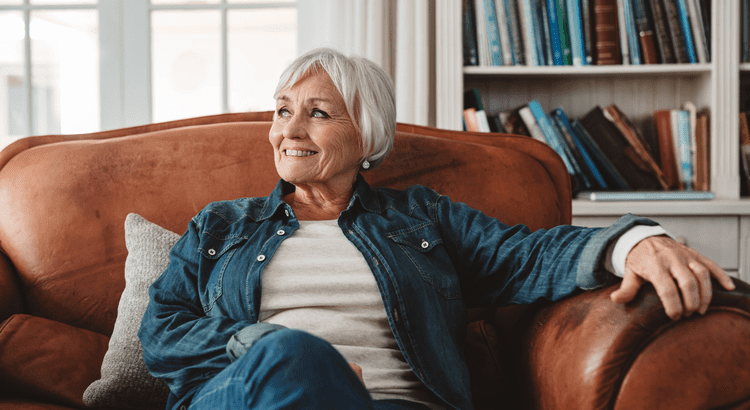 An older woman seated on a couch, surrounded by a bookcase filled with books, exuding a sense of calm and wisdom.