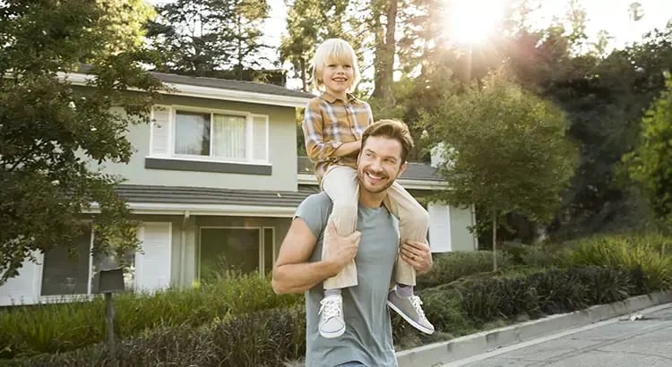 A man giving a child a piggyback ride in front of a house. They are both smiling, indicating a happy family moment.