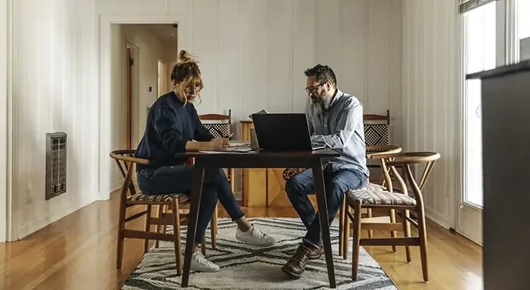 Two people, possibly colleagues, working together at a table with laptops in a modern, well-lit room. The scene suggests collaboration or a business meeting.