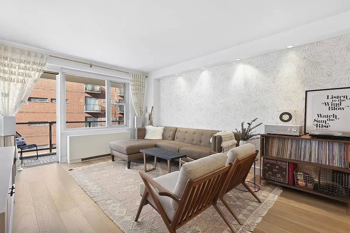 Living room with a taupe sectional, wooden lounge chairs, patterned rug, and windows facing a brick building.