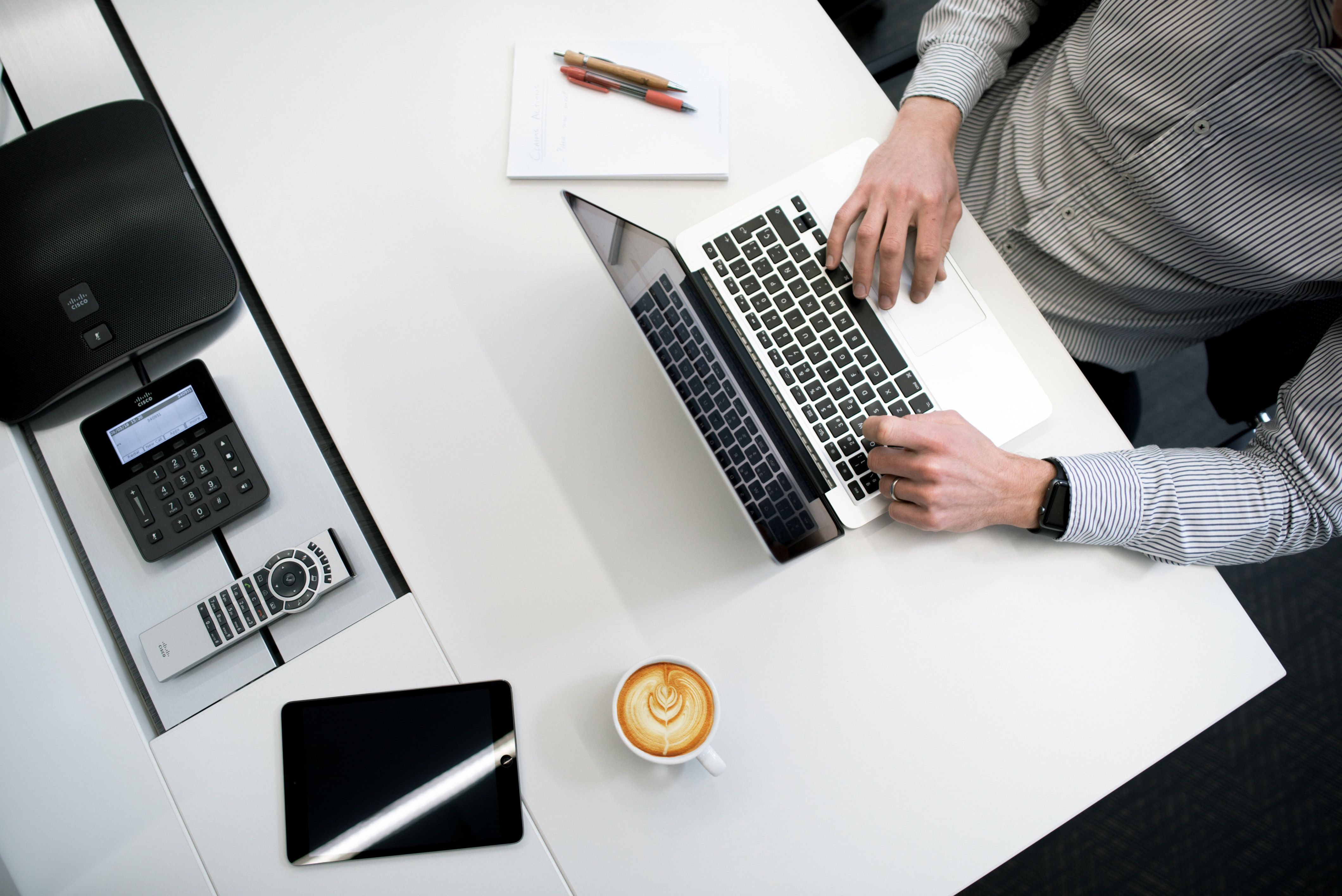 Person typing on a laptop at a desk with a tablet, coffee, and office phone equipment.