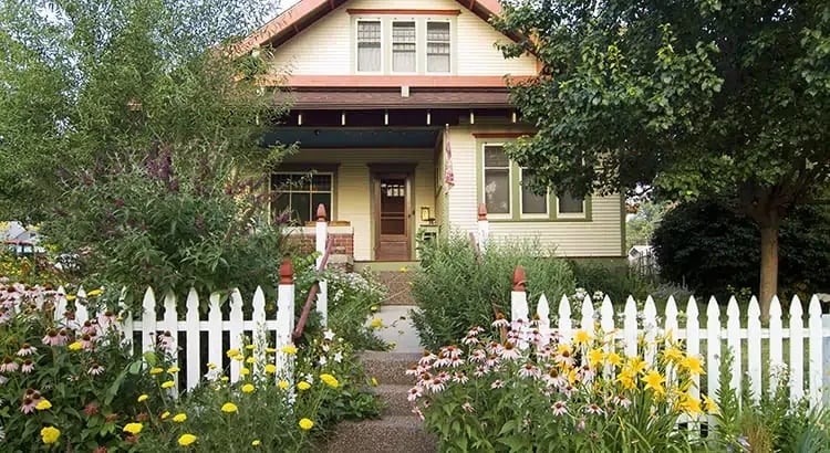 A charming house with a front yard full of flowers and a white picket fence. The house has a welcoming appearance with a porch and greenery.