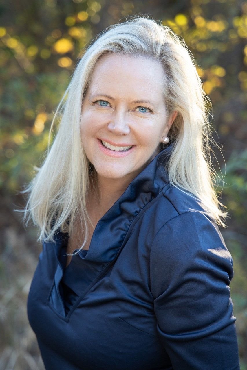 Charlotte Rouse, smiling in a navy jacket against softly blurred foliage.