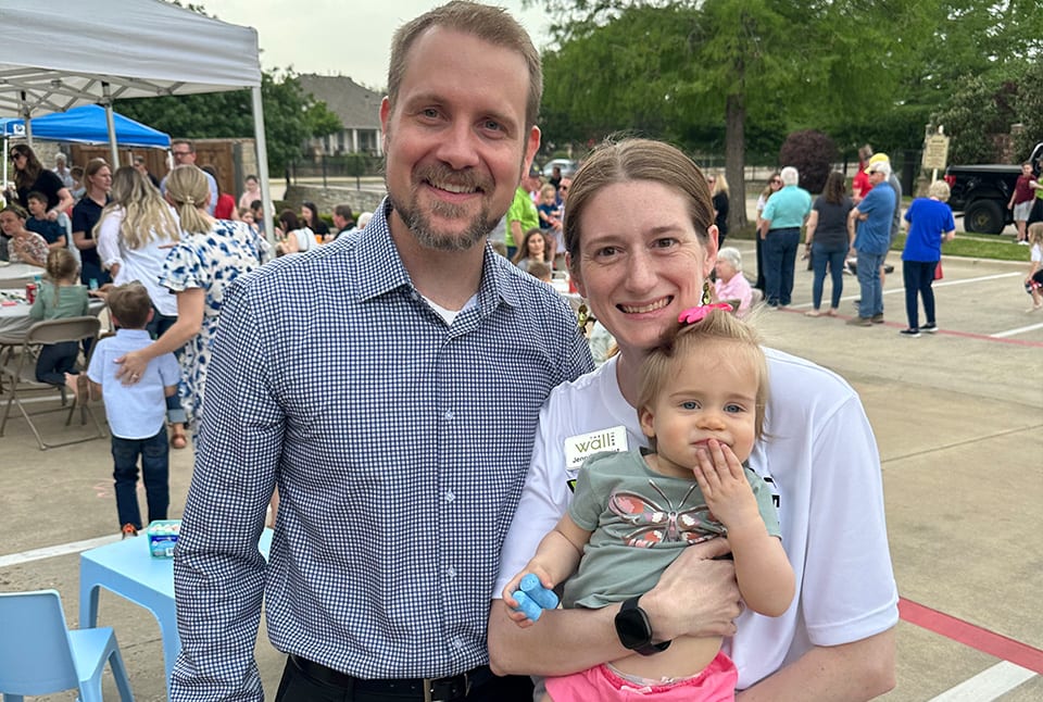 A man in a polo and a woman in a white shirt holding a baby with many people in the background