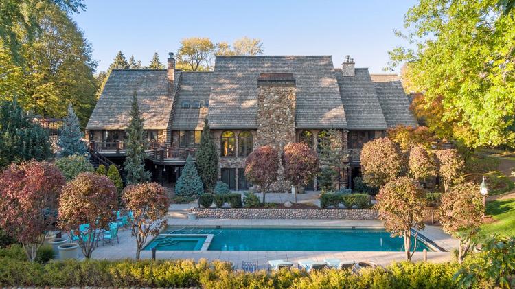 A large, modern brick house with chimneys, a swimming pool surrounded by trees, and a patio.