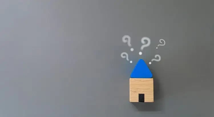 a small wooden house cutout with a blue roof, surrounded by question marks on a light gray background.