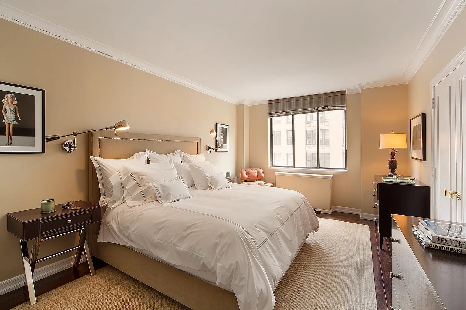 Furnished bedroom with a large white bed, beige headboard, and window overlooking neighboring buildings.