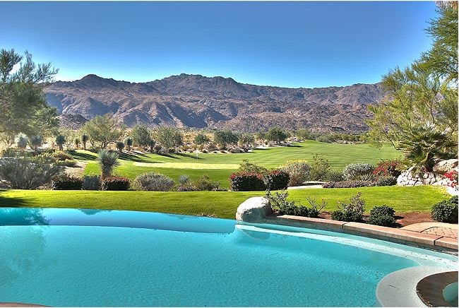 A luxurious swimming pool with a curved edge overlooks a golf course and a mountain range in the background.