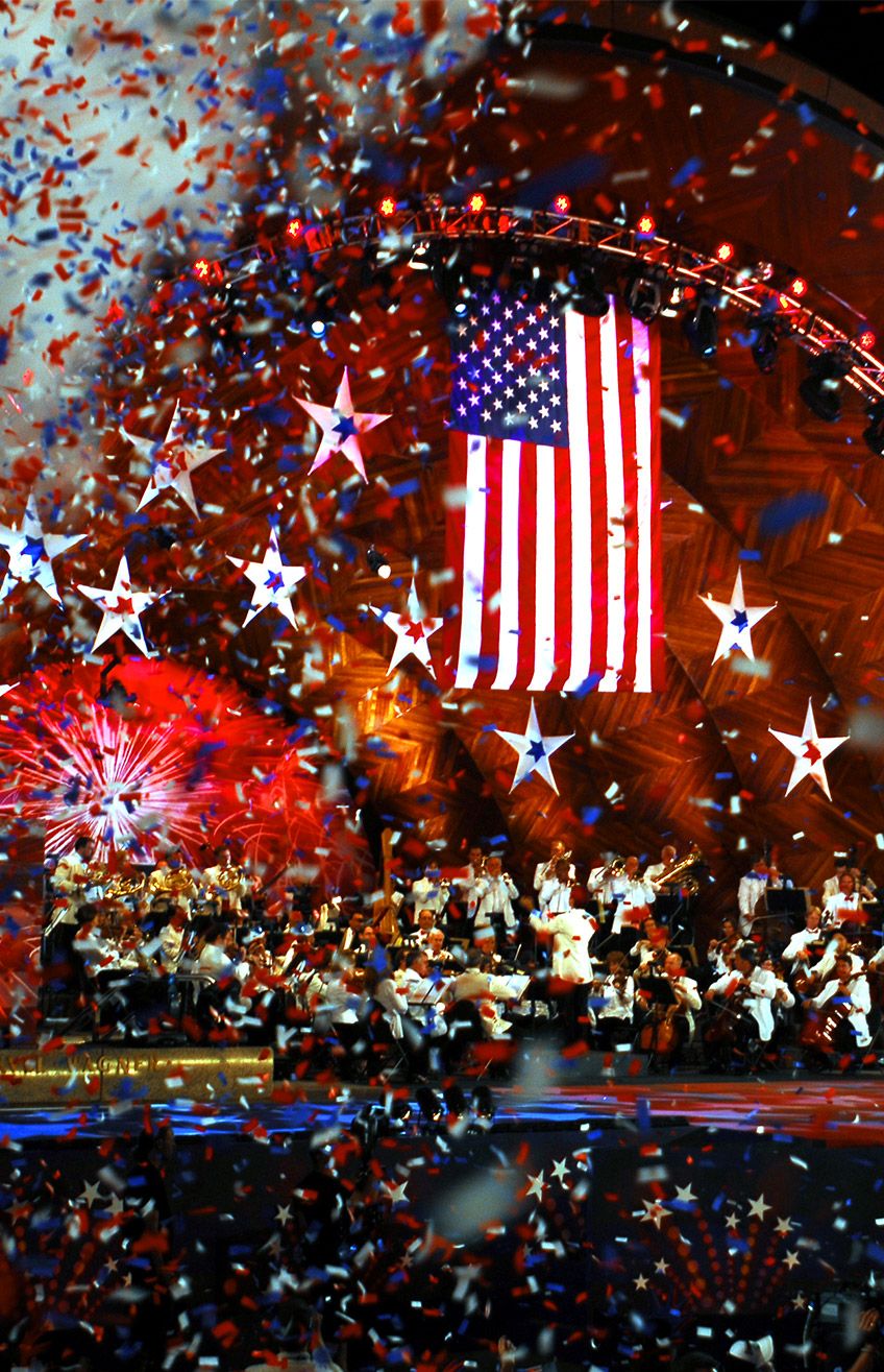 Orchestra performs beneath a large American flag as red, white, and blue confetti falls.