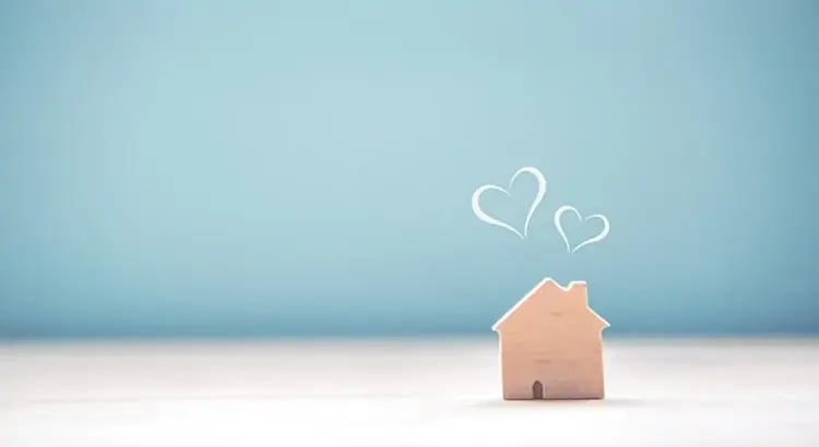 A small wooden house model with heart shapes above it, set against a light blue background. This image likely symbolizes love for home or home ownership.