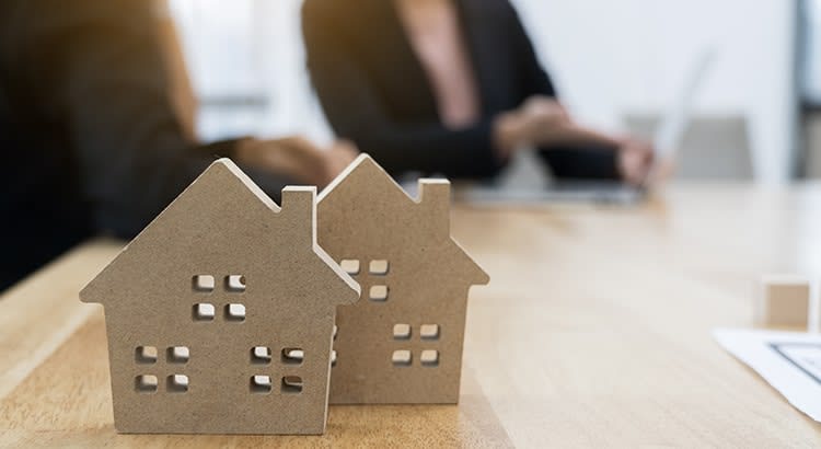 Two small, wooden house models are placed on a wooden table in the foreground, with their windows cut out. In the background, there are blurred figures, suggesting a business meeting or a discussion. 