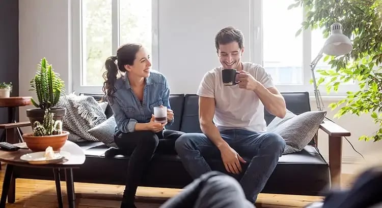 A couple sitting on a couch, engaging in a relaxed conversation and drinking coffee. They seem to be enjoying their time together in a cozy living room setting.