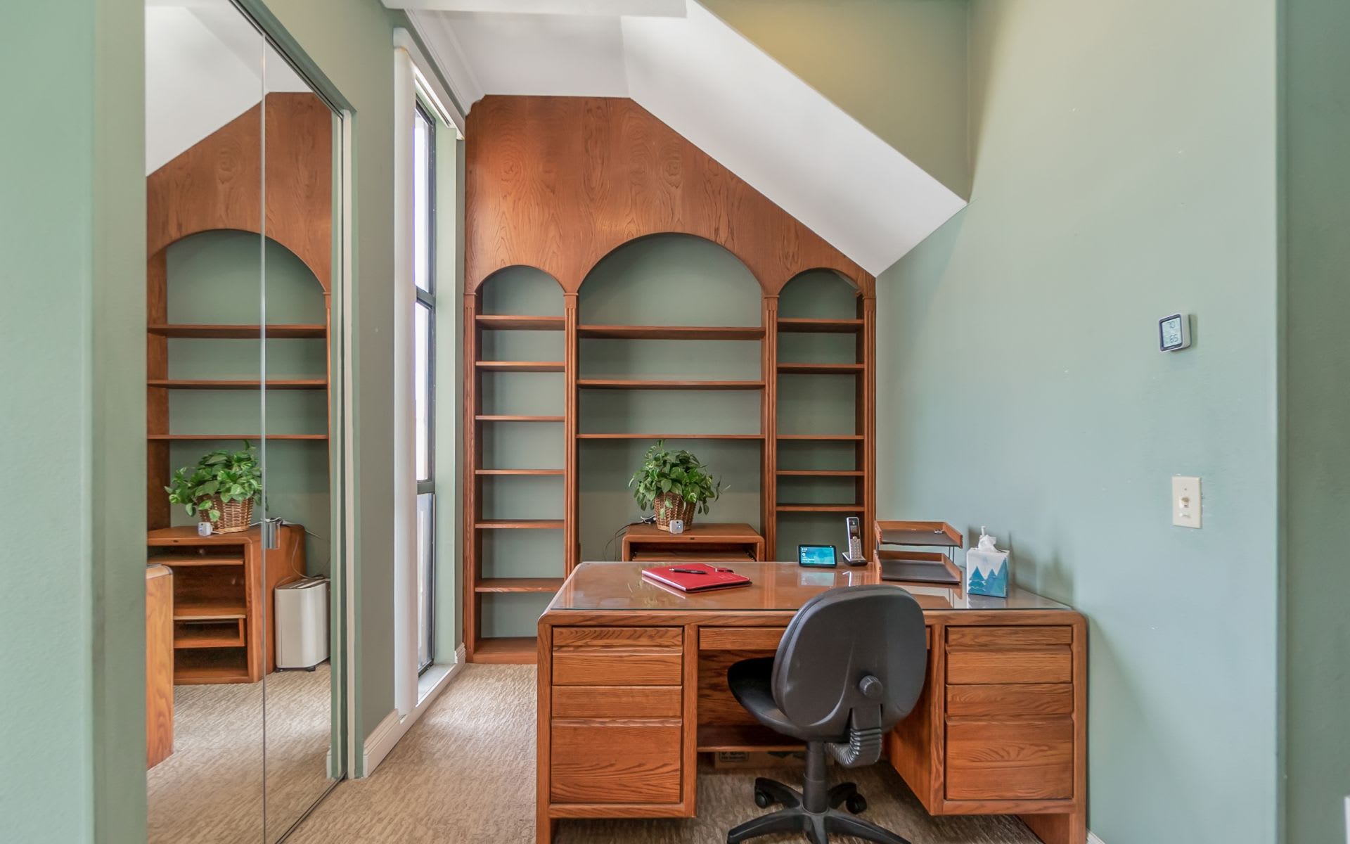 Home office with a wooden desk, arched built-in shelves, mirrored closet door, and sloped ceiling.