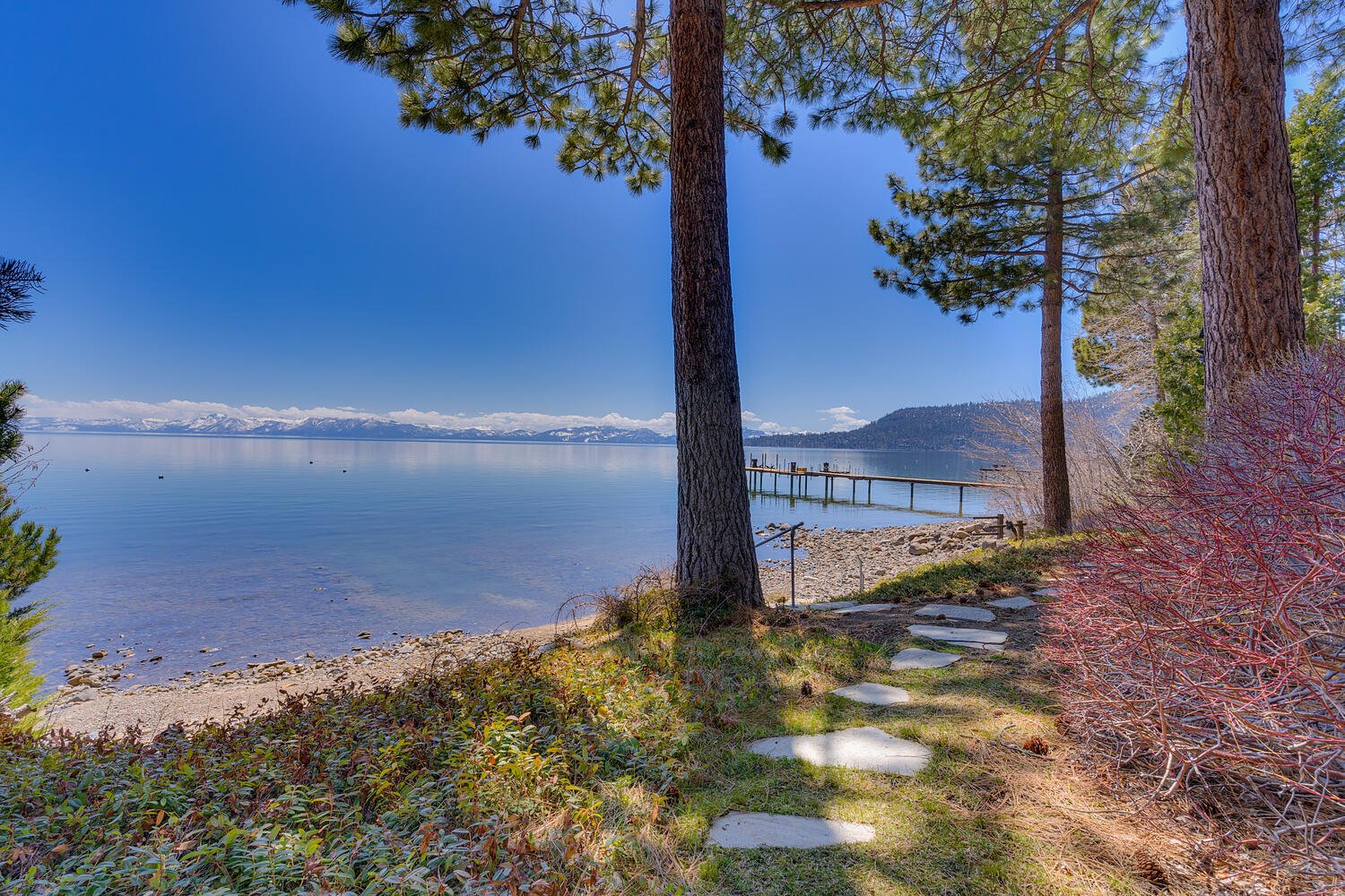 Lake Tahoe viewed from a pine-lined rocky shore, with a pier and distant snow-capped mountains.