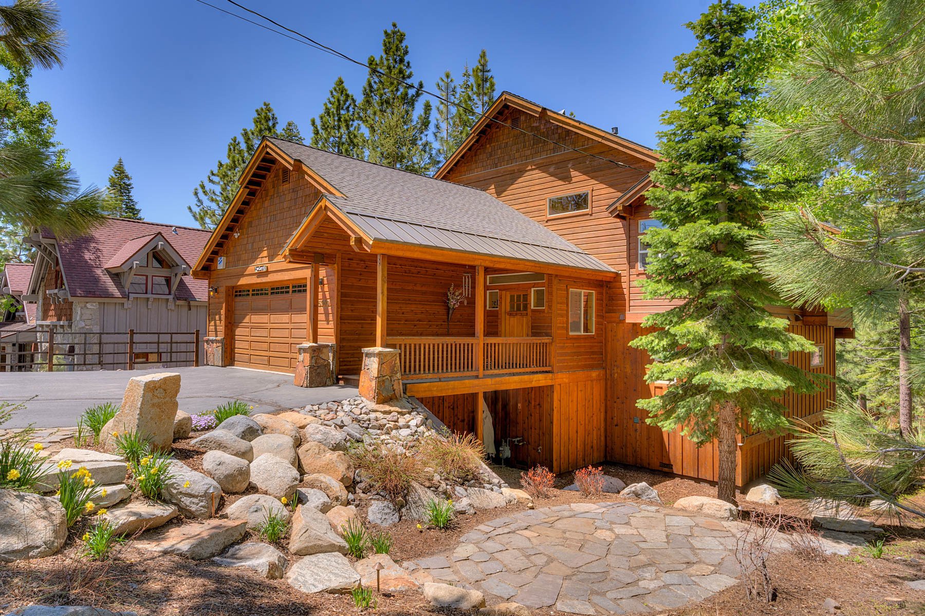 Wood-sided mountain home with a covered porch, garage, and stone landscaping among pine trees.