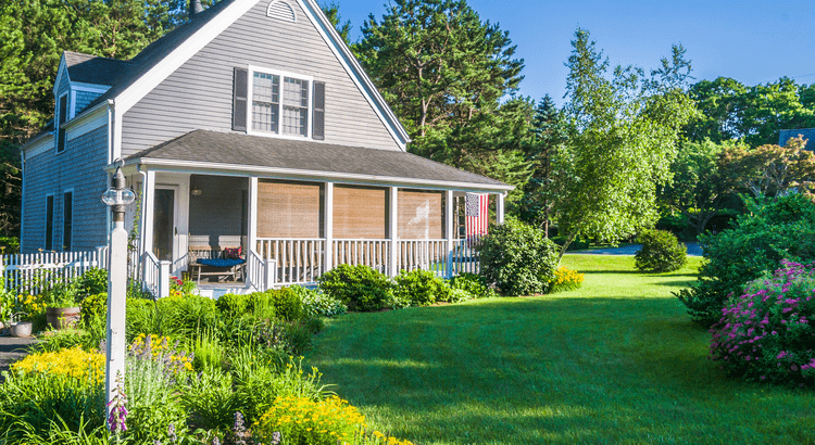Two story house with grey siding and white trim.  Front porch has a white railing and fence.  There are cane type shades hanging from the roof of the porch providing shade.  Additionally there is a US flag hanging from right corner of the porch roof.  It is surrounded by trees plus a large green front lawn.  The lawn has flowing Busches on the right and left sides of the photo. 
