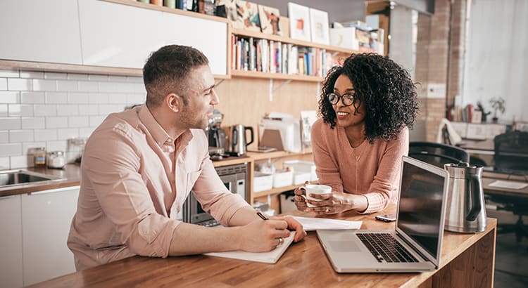 A man and woman appear to be in conversation, with a laptop nearby, suggesting a discussion about finances, home buying, or work.