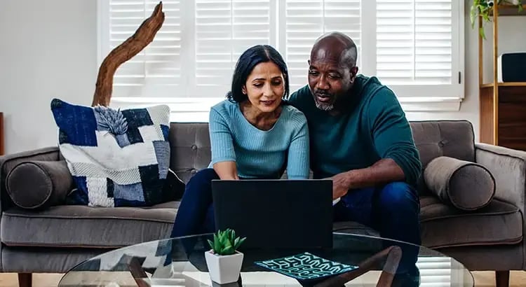 A couple sitting on a couch in their living room, looking at a laptop.