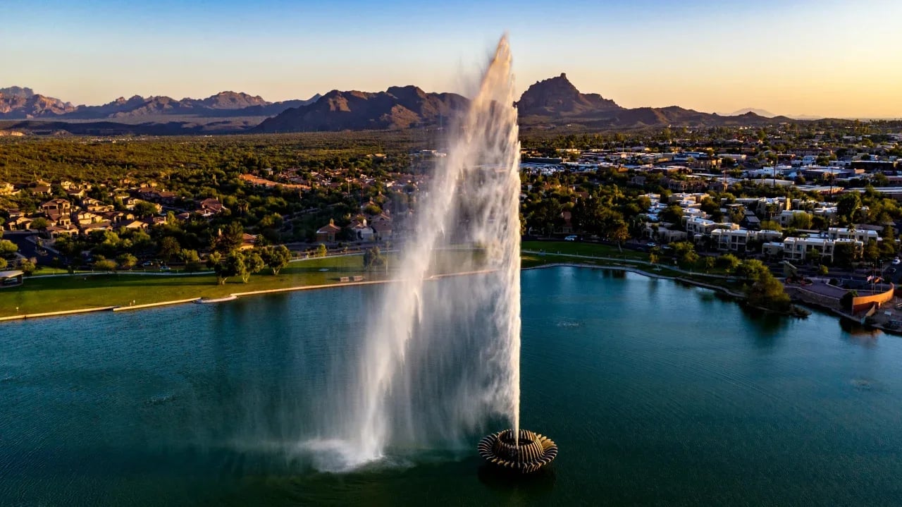 A majestic fountain rises above a lake in a community with luxury homes, bathed in the warm light of sunset.