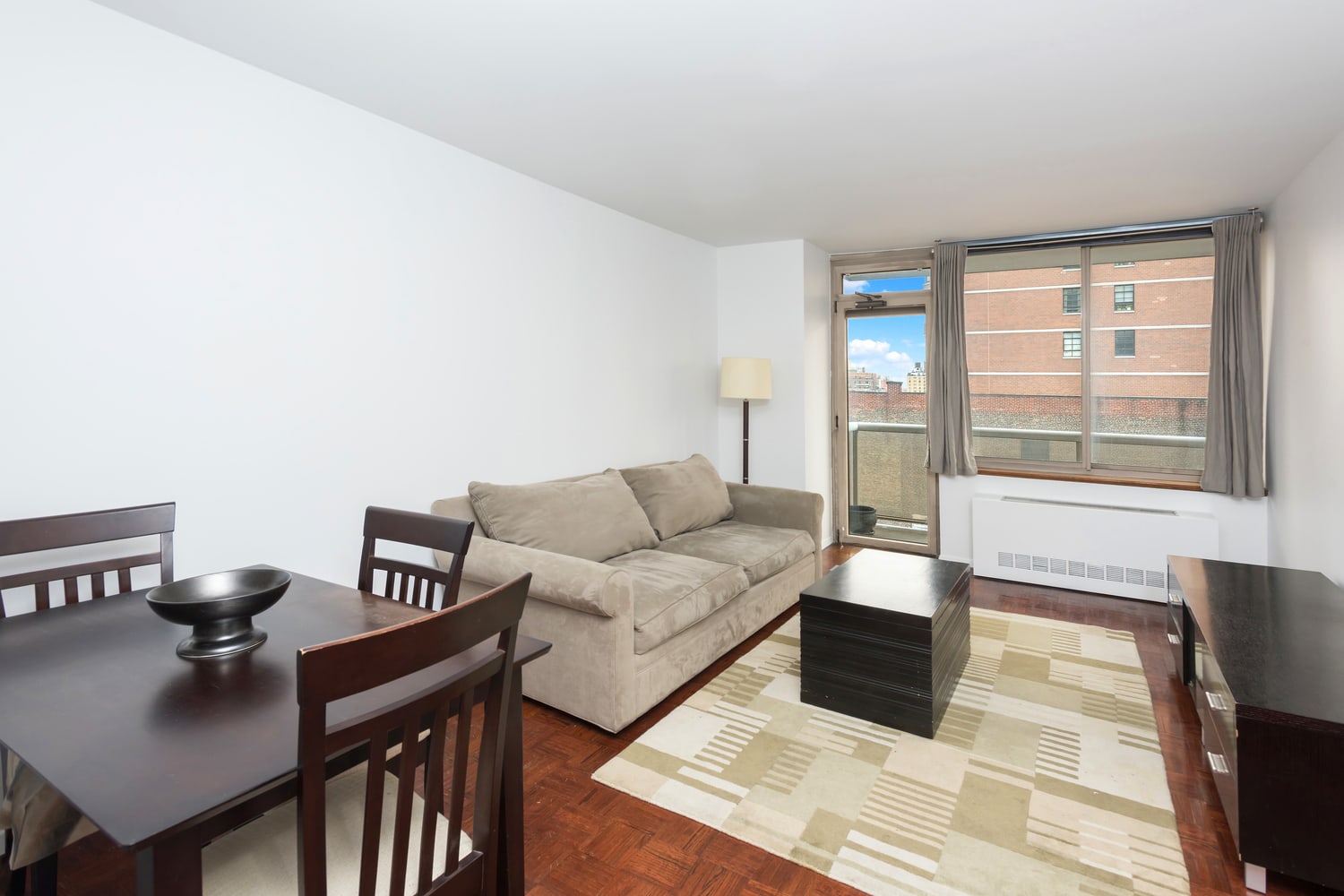 Living and dining area with a beige sofa, dark wood furniture, parquet floors, and a balcony door.
